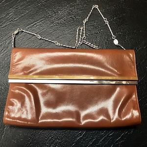 May include: A brown leather clutch bag with a silver chain strap. The bag features a gold-coloured bar across the top and a silver bar below. The rectangular bag has a shiny finish.