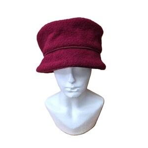 May include: A burgundy cloche hat with a textured, wool-like appearance. The hat features a structured brim and a band detail around the crown. The hat is displayed on a white mannequin head, showcasing its shape and style.