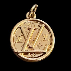May include: A gold-colored pendant with a circular design. The pendant features the letters "LV" in a stylized font, along with floral and geometric patterns. The pendant has a loop at the top for attaching to a chain.