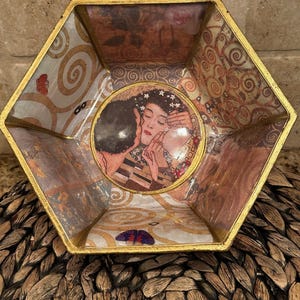 May include: Hexagonal decorative bowl with a gold rim. The interior features a print of Gustav Klimt's "The Kiss" artwork, with swirling patterns and floral accents in shades of brown, gold, and cream.