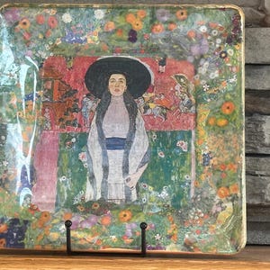 May include: Square decorative tray featuring a painting of a woman in a large black hat. The artwork is surrounded by a floral border with a gold trim. The painting has a red background and green and pink accents.
