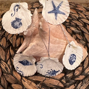 Puede incluir: Adornos de conchas blancas con diseños pintados en azul, incluyendo una estrella de mar y varios patrones de conchas. Cada adorno tiene una cuerda plateada para colgar. Las conchas están dispuestas sobre una superficie tejida.