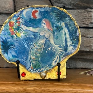 Könnte beinhalten: Eine blaue und weiße Keramik-Muschel mit einem bemalten Meerjungfrauen-Motiv. Die Meerjungfrau hält einen Blumenstrauß und hat rote Haare. Die Muschel steht auf einem goldenen Ständer mit roten und weißen Akzenten.