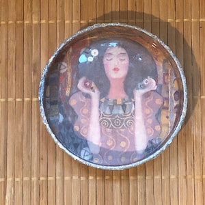 May include: A round, clear glass paperweight with a colorful image of a woman with her eyes closed, holding two small, round objects in her hands. The woman is wearing a purple dress with gold accents.