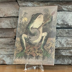 May include: A green frog playing a banjo with a brown wooden body and white strings. The frog is sitting on a brown and gold background with green leaves.
