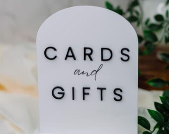 May include: White acrylic sign with black letters that reads "CARDS and GIFTS". The sign is on a white surface with green leaves in the background.