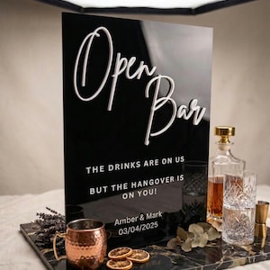 Op de afbeelding: Zwart acryl Open Bar-bord met witte letters. Het bord luidt "Open Bar" en bevat de tekst "THE DRINKS ARE ON US BUT THE HANGOVER IS ON YOU!" en de namen "Amber & Mark" met de datum "03/04/2025".