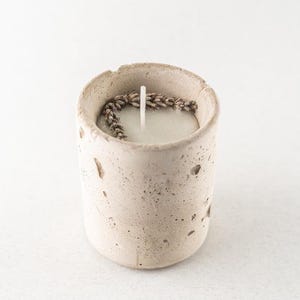 May include: A small, cylindrical candle in a textured, light beige concrete holder. The candle is pale green with a white wick. Dried lavender sprigs encircle the candle's top edge, adding a natural touch to this home decor item.