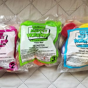 May include: Three sealed Happy Meal toys from the Jim Henson's Muppet Workshop series. The packages are labeled "Dog", "Monster", and "What-Not", each containing a figure and two accessories. The packages are in pink, green, and blue.