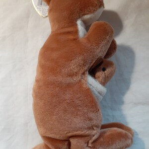 May include: A brown plush kangaroo holding a smaller brown plush joey in its pouch. The kangaroo is standing on a white surface.