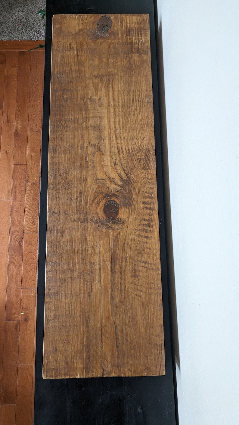 May include: A close-up of a wooden shelf with a natural wood finish. The shelf has a knot in the center and is mounted on a black wall.