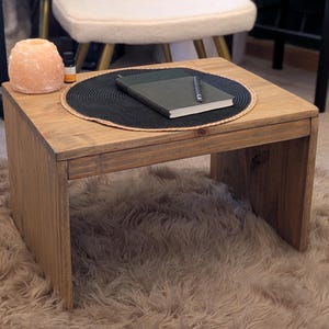 May include: A small, square wooden coffee table with a natural finish. A black placemat holds a green notebook and a pen. The table sits on a fluffy, light brown rug. A Himalayan salt lamp and essential oil bottle are visible.