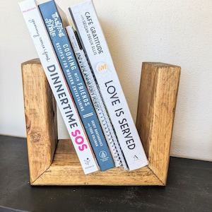 May include: A wooden bookend shaped like the letter U, holding a stack of cookbooks. The books are titled "Love is Served", "Anti-inflammatory Foods", "Cooking with Friends", "Dinnertime SOS", and "Tasting Table Recipes".