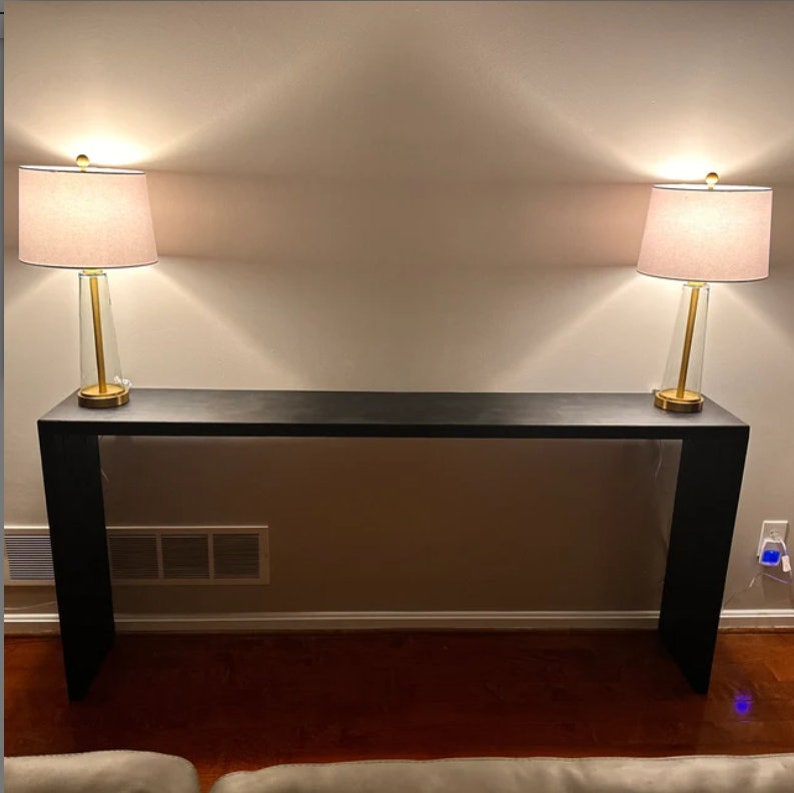 May include: A black console table with two lamps on top. The lamps have gold bases and white lampshades.