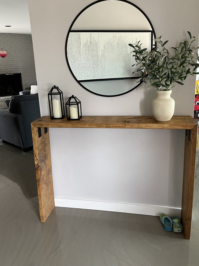 May include: A rustic wooden console table with a natural wood finish. The table has a black metal frame and is decorated with two black metal lanterns and a vase with green foliage.