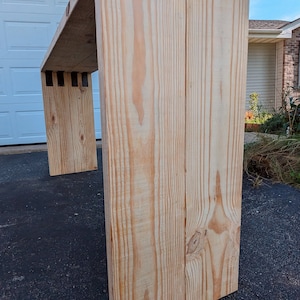 May include: A light wood table with a simple design. The table is made of natural wood and has a light brown finish. The table is shown from the side, with the legs visible. The table is sitting on a black surface.