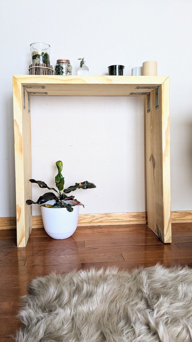May include: A wooden console table with a light wood finish. The table has a shelf and is decorated with small plants and jars. A potted plant sits on the floor in front of the table.
