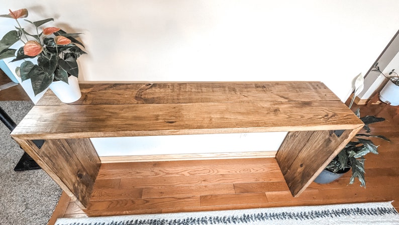 May include: A rustic wooden console table with a natural wood finish. The table has a rectangular top and two open shelves. The table is made of reclaimed wood and has a distressed look.