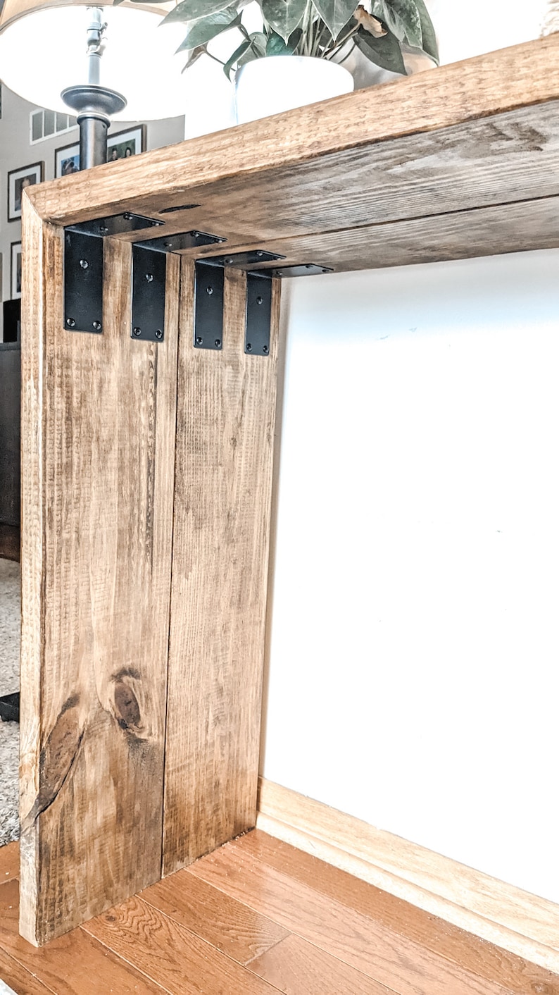 May include: A wooden console table with a natural wood finish and black metal brackets. The table is shown in a room with hardwood floors.