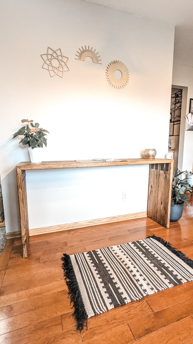 May include: A wooden console table with a natural wood finish and black metal accents. The table has a rectangular top and two legs. There is a black and white striped rug on the floor in front of the table. There are three wooden wall decorations above the table, a sun, a star, and a circle.