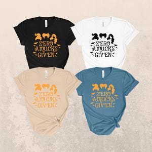 May include: Four short-sleeve t-shirts in black, white, beige, and blue. Each shirt features a graphic of three stylized hairstyles above the text "ZERO AMUCKS GIVEN" and small bat silhouettes. The designs are printed in contrasting colors.