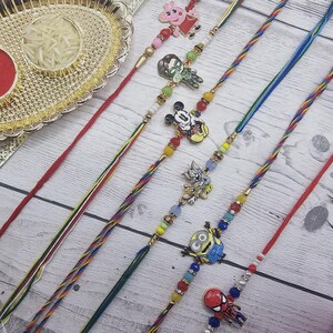 May include: A selection of vibrant Rakhi bracelets featuring cartoon character charms, beads, and twisted cords. A decorative gold tray holds red powder and rice. A peacock feather and rose are in the background.