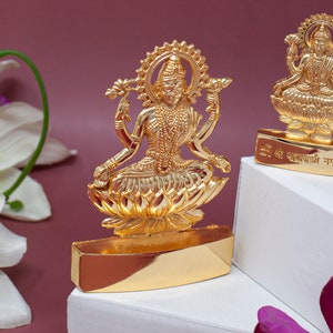 May include: Two gold-coloured statues of the Hindu goddess Lakshmi, each sitting on a lotus flower. The statues are on white bases with the text "श्री लक्ष्मी नारायण" in Hindi.