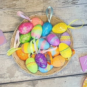 May include: A wicker basket filled with colourful glittery Easter eggs with ribbons attached. The basket is on a wooden surface with two signs that say "Egg Hunt This Way" and "That Way".