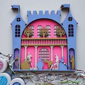 Puede incluir: Un castillo de madera cortado con láser en azul y rosa, con balcón, ventanas arqueadas y siluetas de figuras con vestimenta de época. Candelabros dorados y banderas adornan la estructura. Pieza de arte decorativa.