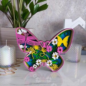 Peut inclure: Une d&eacute;coupe de papillon en bois multicouche pr&eacute;sentant une libellule rose, un papillon jaune et diverses fleurs blanches et roses sur un fond bleu. Le design utilise des couches superpos&eacute;es pour cr&eacute;er de la profondeur, avec une bordure rose.