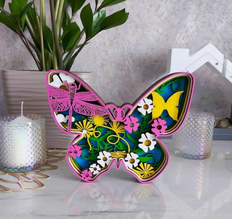 Peut inclure: Une d&eacute;coupe de papillon en bois multicouche pr&eacute;sentant une libellule rose, un papillon jaune et diverses fleurs blanches et roses sur un fond bleu. Le design utilise des couches superpos&eacute;es pour cr&eacute;er de la profondeur, avec une bordure rose.