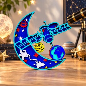 May include: A layered wooden crescent moon decoration featuring a blue background with white stars, a red UFO, a yellow planet, a white astronaut, and a white space shuttle. A blue and cyan satellite and a globe are attached to the front of the moon.
