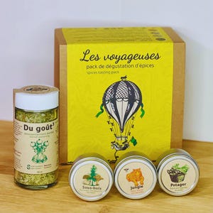 Op de afbeelding: Een set van vier specerijenpotten met een ontwerp van een luchtballon op de doos. De potten zijn gelabeld met "Du goût", "Sous-bois", "Jungle" en "Potager". De doos is gelabeld met "Les voyageuses pack de dégustation d'épices spices tasting pack".