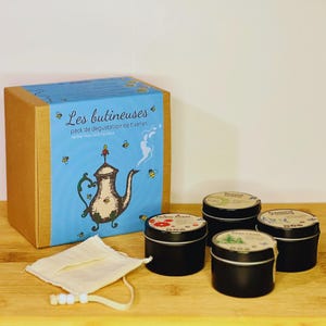 Peut inclure: Une boîte brune avec une illustration bleue et blanche d'une théière et d'abeilles. La boîte indique "Les butineuses pack de dégustation de tisanes Herbal teas tasting pack". Il y a quatre petites boîtes noires avec des étiquettes colorées et un sachet de thé en mousseline blanche.