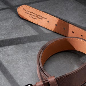 May include: A brown leather belt with a dark metal buckle. The inside of the belt is light brown and features an engraved message: 'Now you will always think of me when you take off pants! Love you!' The belt is shown on a gray textured surface.