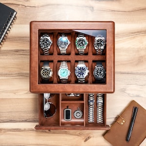 May include: A brown leather watch display case with a glass top, showcasing eight wristwatches with various face designs and metal bands. The bottom drawer holds sunglasses, pendants, and watch straps. A notebook and pen are visible.
