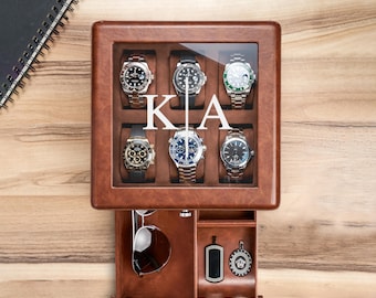 May include: A brown leather watch box with a glass lid featuring the white letters KA. The box contains six wristwatches in individual compartments. A pull-out drawer below holds a pair of sunglasses, a rectangular dog tag, and a round pendant.