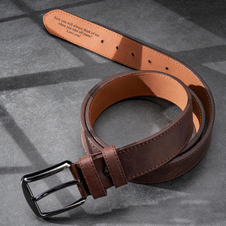 May include: A brown leather belt with a dark metal buckle. The inside of the belt is light brown and features an engraved message: 'Now you will always think of me when you take off pants! Love you!' The belt is shown on a gray textured surface.