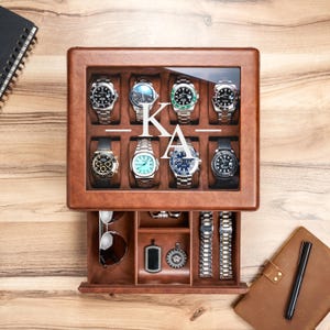 May include: A brown leather watch box with a glass lid featuring the white monogram KA. The top section holds eight wristwatches in individual compartments. The bottom pull-out drawer contains sunglasses, two metal watch bands, and two pendants.