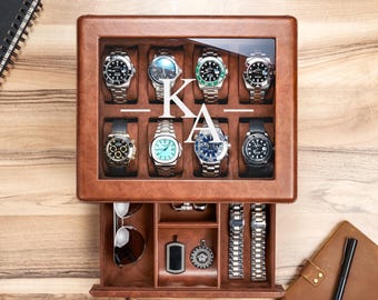 Puede incluir: Una caja de cuero marrón para relojes con tapa de cristal que presenta el monograma blanco KA. La sección superior contiene ocho relojes de pulsera en compartimentos individuales. El cajón inferior contiene gafas y dos correas metálicas.