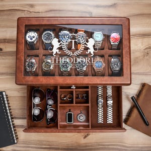 May include: A brown leather watch box with a glass lid featuring the text THEODORE and a lion crest. The top section holds twelve wristwatches, while the bottom drawer contains two pairs of sunglasses, two rings, cufflinks, two pendants, and two watch bands.