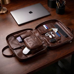 May include: Open brown leather briefcase with multiple compartments, including a small pouch and a section for a phone. The briefcase contains a wallet, keys, and other accessories. A laptop is visible in the background.