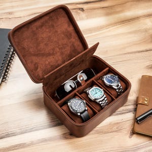 May include: A brown leather watch case with a zippered closure, open to reveal three watches and a pair of sunglasses. The interior is lined with a soft, brown material. The watches have silver and black bands.
