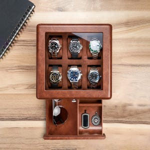 May include: A brown leather watch display case with a glass top, showcasing six wristwatches with various band and face designs. A drawer below holds sunglasses and two pendants. The case sits on a wooden surface.