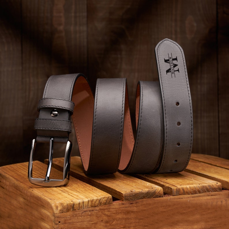 Pu&ograve; includere: Una cintura in pelle grigia scura con una fibbia argentata e il nome "Matthew" inciso sul cinturino.