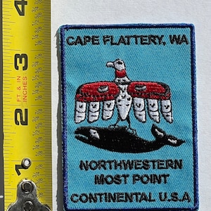 May include: A rectangular patch with a blue background and a dark blue border. The patch features an eagle and a whale design, with text that reads "CAPE FLATTERY, WA", "NORTHWESTERN MOST POINT", and "CONTINENTAL U.S.A.". The patch measures approximately 3 inches by 2 inches.