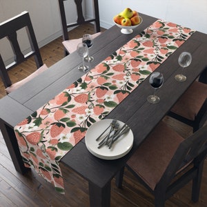 May include: A pink and green table runner with a floral and strawberry pattern. The runner is on a dark wood table with two chairs and a bowl of fruit.