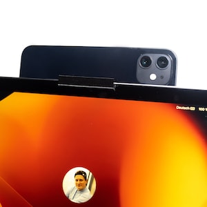 Pode incluir: Um tablet preto com uma tela laranja brilhante exibindo uma videochamada com uma pessoa. O tablet está posicionado em cima de um smartphone preto com uma lente de câmera dupla.