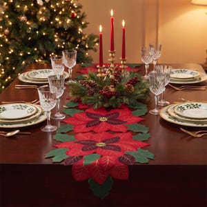 May include: A red and green poinsettia-shaped table runner featuring layered fabric petals and leaves. The runner is centered on a dark wood table with white plates, clear crystal glasses, and a gold candelabra holding three red taper candles.