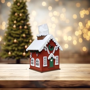 May include: A red and white gingerbread house shaped tissue box cover. The house has a white roof with a candy cane trim and a green wreath on the front. The tissue box cover is sitting on a wooden table.